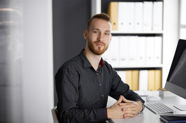 Red-bearded friendly adult businessman looking at camera. Business headshot or portrait in office.