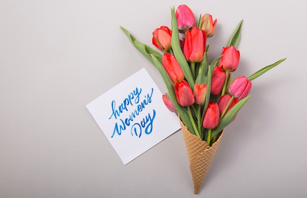 Red  beautiful tulips in an ice cream waffle cone with card Womans Day on a concrete background. Conceptual idea of a flower gift. Spring mood