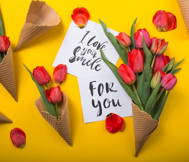 Red beautiful tulips in an ice cream waffle cone with ink card on a yellow color background. conceptual idea of a flower gift. spring mood