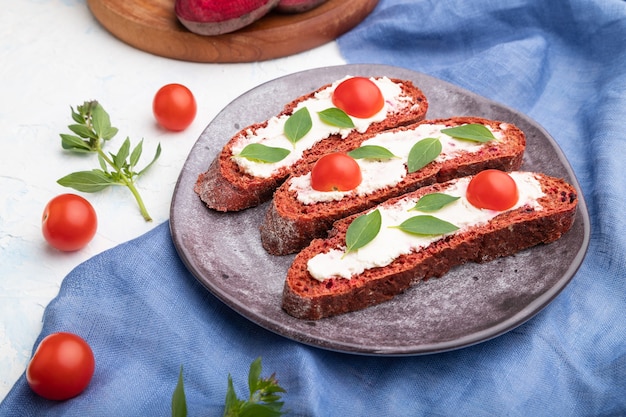 Red beet bread sandwiches with cream cheese and tomatoes on white concrete surface and blue linen textile