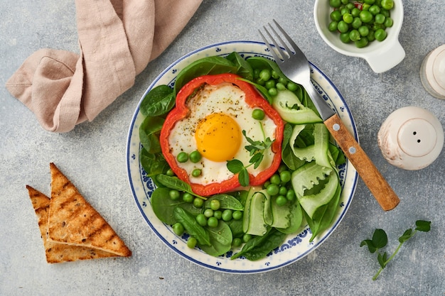 明るい灰色のテーブルの背景に朝食プレートに卵、ほうれん草の葉、グリーンピース、マイクログリーンを詰めた赤ピーマン。上面図。