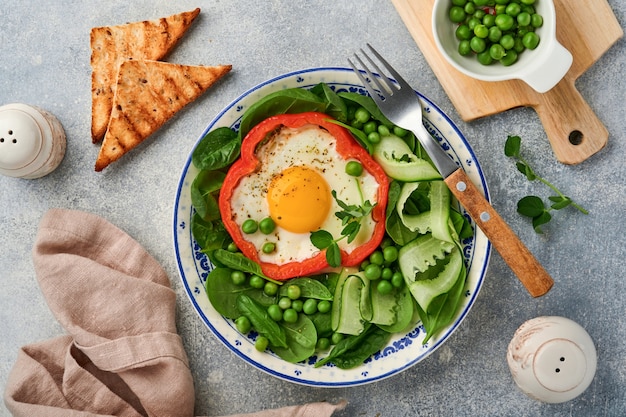 Красный сладкий перец, фаршированный яйцами, листьями шпината, зеленым горошком и микрозеленью на тарелке для завтрака на светло-сером фоне стола. Вид сверху.