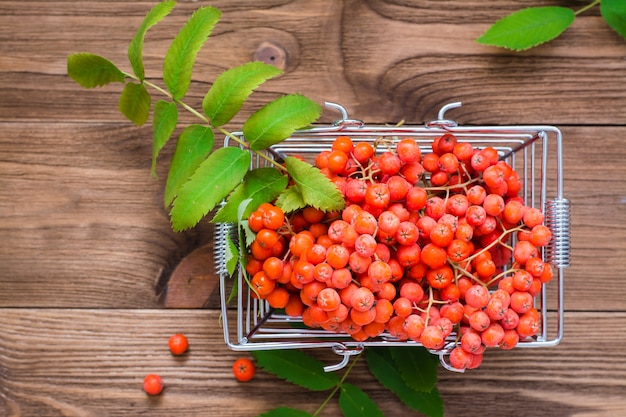 Red berries and rowan leaves in a metal basket on woodtable Top view