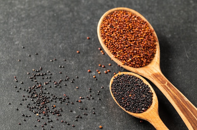 Semi rossi e neri della quinoa in cucchiai di legno su fondo strutturato nero