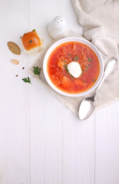 Minestra rossa del borscht in ciotola bianca con panna acida e prezzemolo, vista superiore, su fondo bianco di legno.