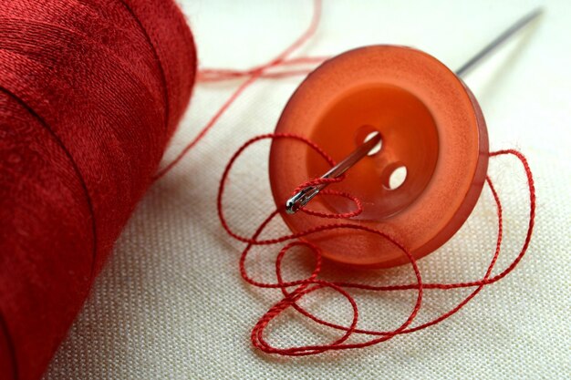 Красная кнопка с иглой со вставленной нитью на белой льняной ткани макросъемка крупным планом