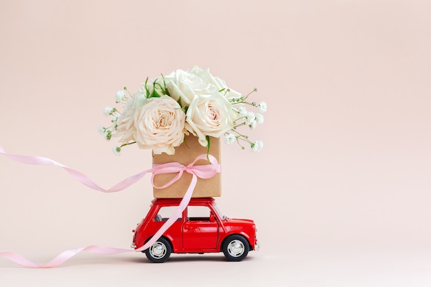 Automobile rossa con una confezione regalo di fiori di rose sul tetto su sfondo rosa. buon san valentino, festa della mamma, 8 marzo, concetto di biglietto di auguri per la giornata mondiale della donna, consegna di fiori.