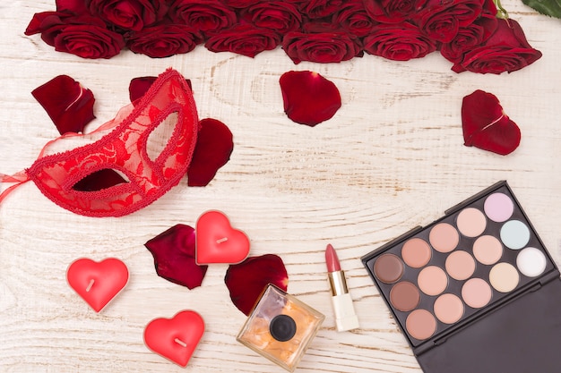 Red carnival mask, bouquet of red roses, lipstick, bottle of perfume and eye shadow on light wooden background.