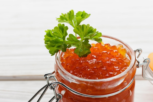 Red caviar in jar with fresh parsley Seafood snack on white wooden table Salmon delicacy close up Side view