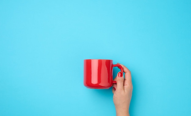 青い背景の上の女性の手に赤いセラミックカップ、飲み物と手を上げる、コーヒーブレイク