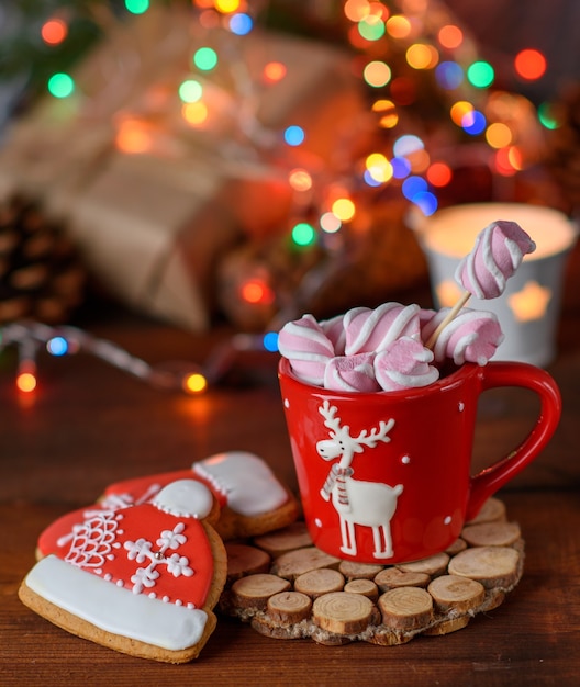 Red ceramic cup with cocoa and marshmallows and baked gingerbread, Christmas lights behind