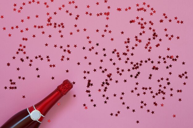 Red Champagne bottle with confetti stars on pink background. Copy space, top view