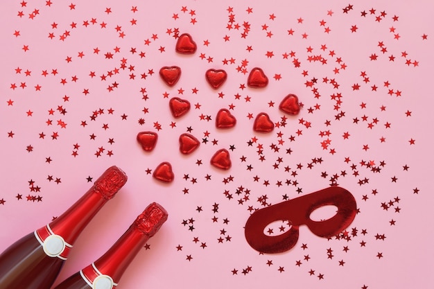 Красные бутылки шампанского с сердечками из красного шоколада и карнавальной маской на розовом. Копировать пространство