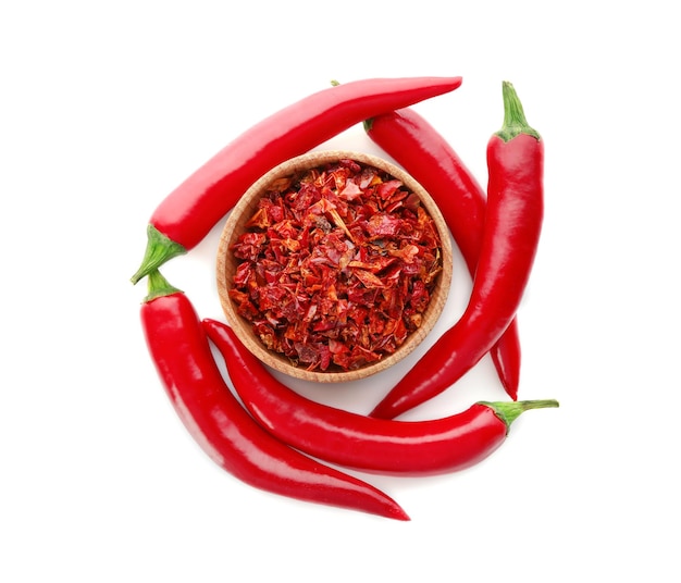 Red chili flakes in wooden bowl with fresh pepper pods on white background