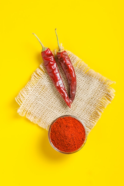 Red chilli powder in glass bowl and chilli peppers on yellow surface