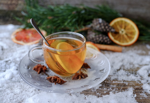 Red Chinese lemon tea in glass Cup on snow-covered wooden table with pine branches, cones, anise, dried oranges