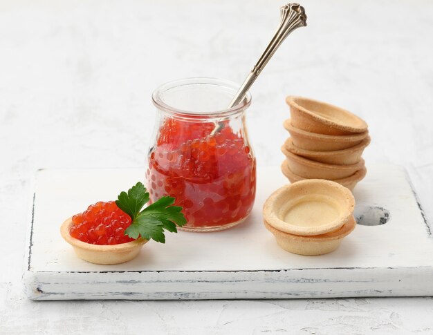 Photo red chinook caviar in a glass jar and round tartlets on a white table