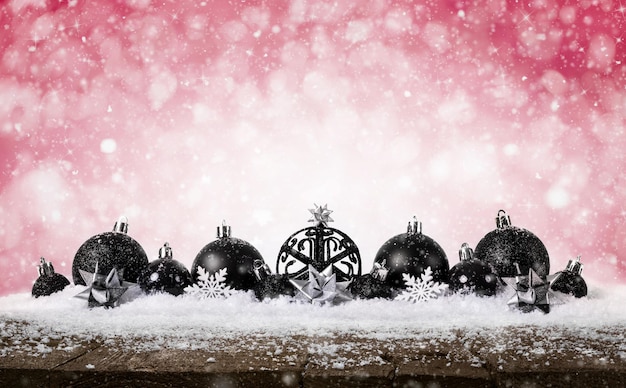 Красный новогодний фон - украшенные черные шары на снегу со снежинками и звездами на деревянном столе.