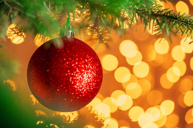 Red christmas ball hanging on fir tree branch over golden bokeh lights 