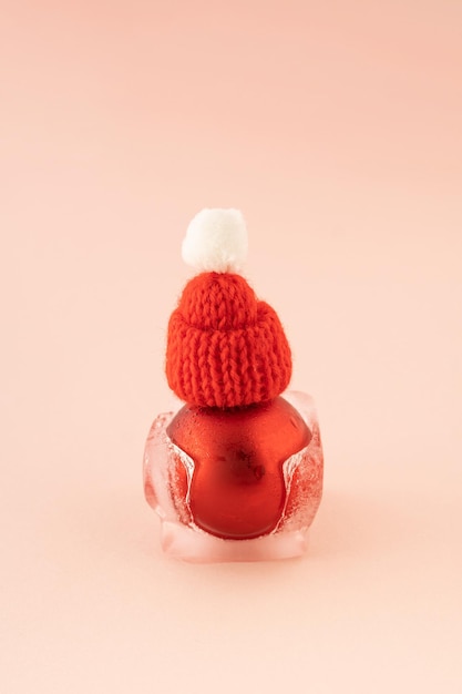 Red Christmas bauble with Santa's hat in melting ice cube on pink background New Year greeting card