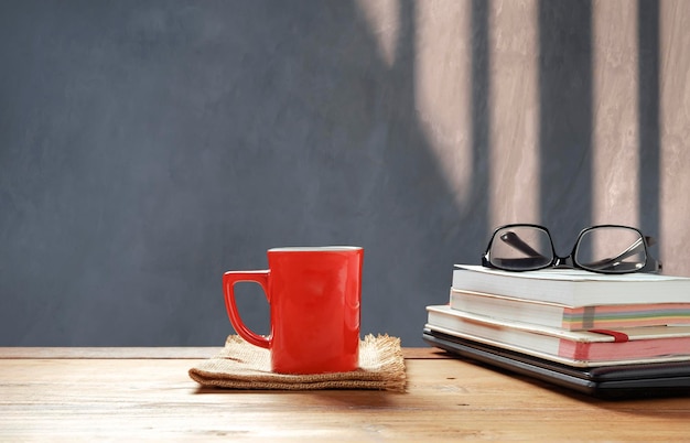Red coffee cup and eyeglasses on stacked books with black laptop on wooden table in loft living room