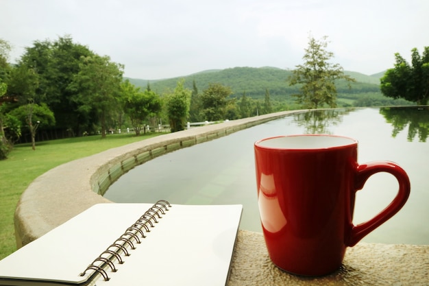 Una tazza di caffè rossa e un taccuino sono posizionati sul bordo della piscina in un cortile, con vista sulle montagne.