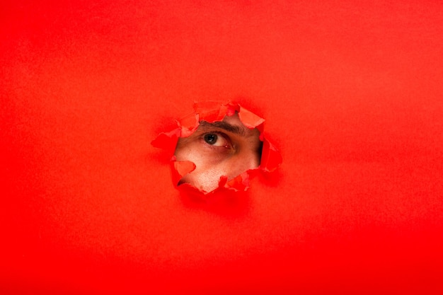 Red color paper with a ripped hole and watching eye from it, the concept of rumors and peeping. Funny picture of a woman's eye looking for interesting projects.