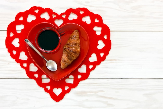Foto tazza rossa di tè nero e croissant freschi fatti in casa sul piatto di forma cuore rosso.