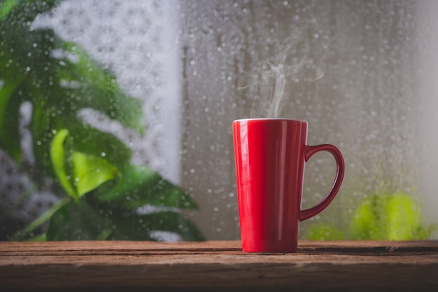 Red cup of coffee beside window and the rain on tropical plant