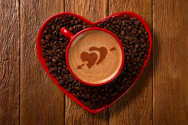 Red cup and coffee saucer in heart shape with decorated coffee on old wood space. Top View. Heart shape in coffee.