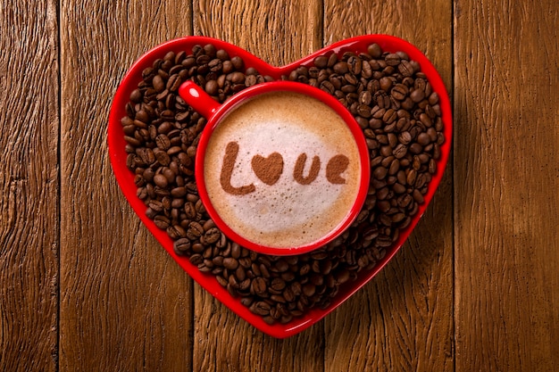 Red cup and coffee saucer in heart shape with decorated coffee on old wood space. Top View. Written love shape in coffee in english.