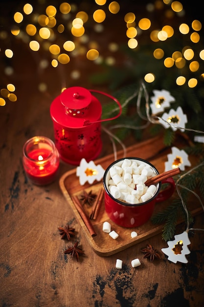 Красная чашка горячего какао с зефиром на деревянном столе с елкой и светящейся гирляндой для ...