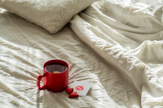 Tazza di tè rossa e due cuori di biscotti, una nota con un bacio su un letto bianco