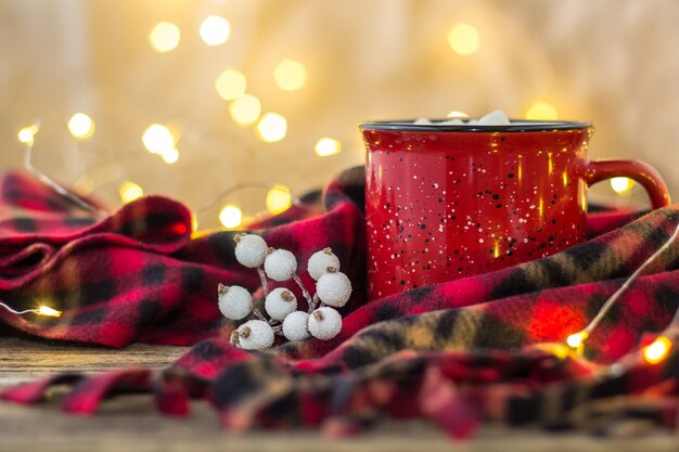 Red cup with hot coffee drink and sweet marshmallows on wooden table with plaid in Christmas decorations with berries and garland bokeh lights background. New Year holidays concept.