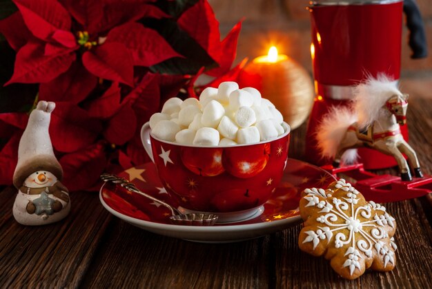 Red cup with hot drink and marshmallow with gingerbread. Christmas concept with poinsettia flower, burning candle and christmas decor. Close-up, selective focus, shallow depth of field