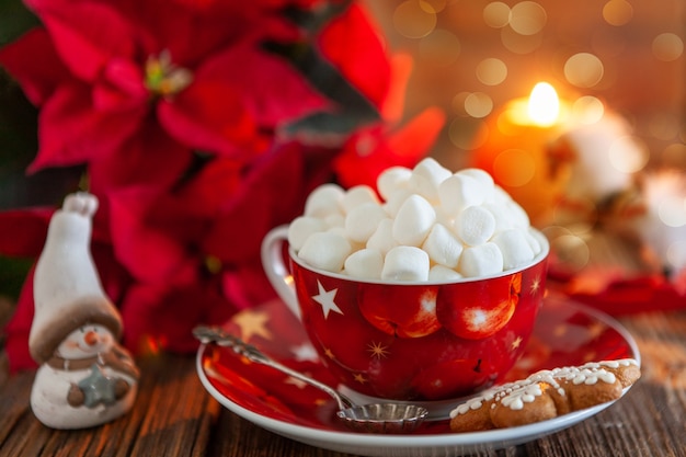 Red cup with hot drink and marshmallow with gingerbread. Christmas concept with poinsettia flower, burning candle and christmas decor. Close-up, selective focus, shallow depth of field