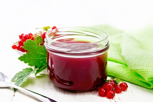 Red currant jam in a glass jar, bunches of berries with leaves, a towel and a spoon on wooden board background