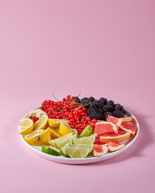 Красная смородина, шелковица и кусочки лимона, лайма, грейпфрута в тарелке на розовом фоне с копией пространства. Набор для приготовления летнего витаминного напитка