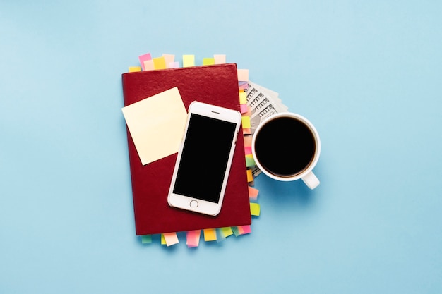 Red diary with stickers on the pages, a cup with black coffee, one hundred dollars, white phone, blue background. Concept of a successful business, proper planning, time management. Flat lay, top view