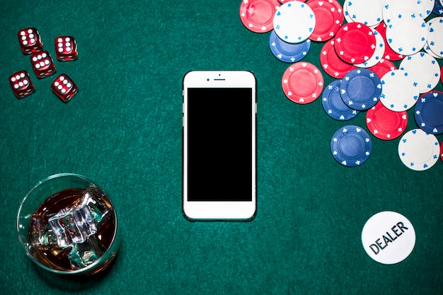 Фото Красные кубики; стекло виски; чипы казино; чип дилера и сотовый телефон на покерном столе