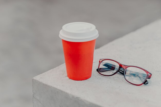 Red disposable cup of hot beverage on white concrete wall, optical glasses near. 