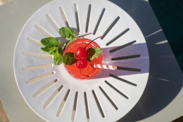 Photo red drink with peppermint leaves and cherries in a  glass with carton straw on the shores of a pool