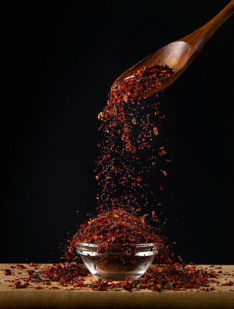 Red dry fragrant spices are poured into a glass salt cellar from a wooden spoon Place for advertising on a black background