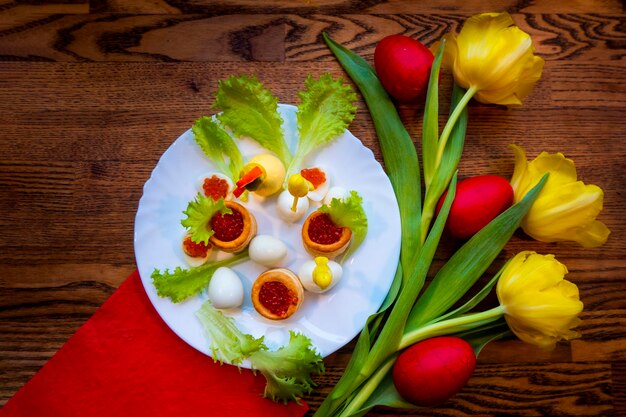 Красные яйца лежат на столе среди желтых тюльпанов, привязанных к красной ленте. Тарелка с яйцами перепелок, гнездами для икры и курицей из желтков - это концепция пасхального блюда.