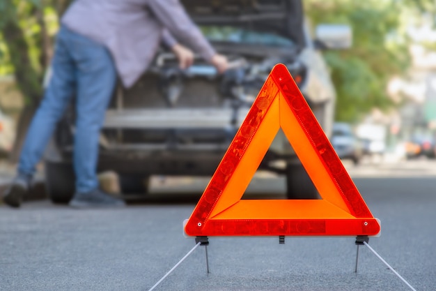 Red emergency stop triangle sign afore Destroyed car in car crash traffic accident on city road. Man driver looking on Smashed broken car in accident. Copy space.