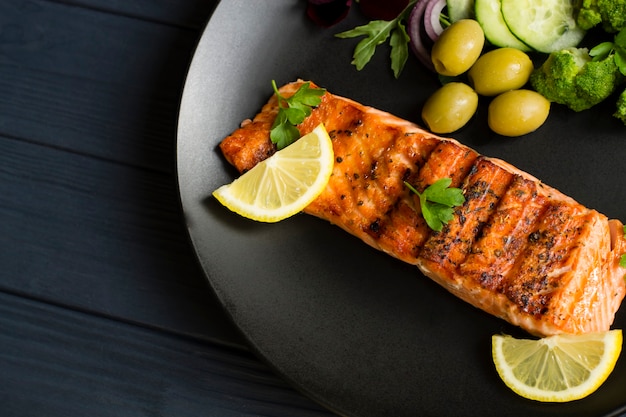 Red fish steak, on a black plate. With lemon and olives. Top view.