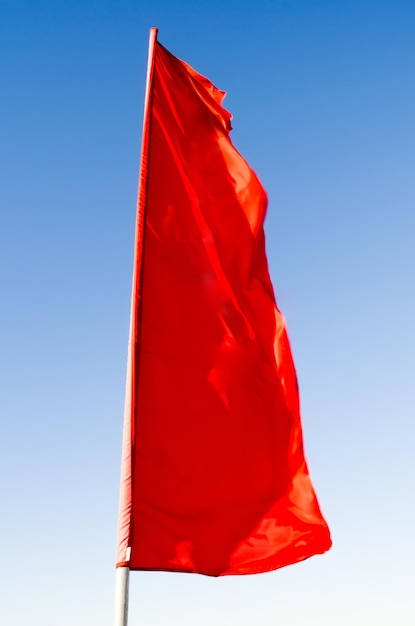 Red flag of rectangular shape elongated fluttering in the wind on the flagpole background of the blue sky