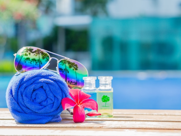 Red flowers, sunglasses, shampoo, lotion and rolled up towels at the side of swimming pool