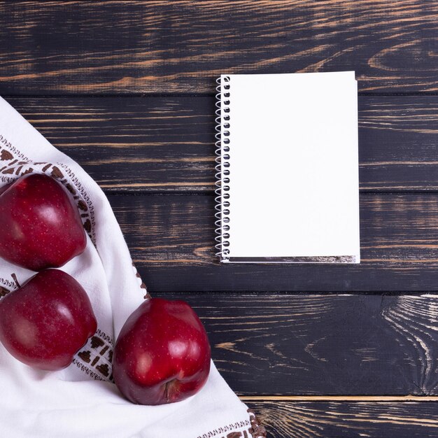 Красные свежие яблоки на темном деревянном фоне с полотенцем и белым блокнотом