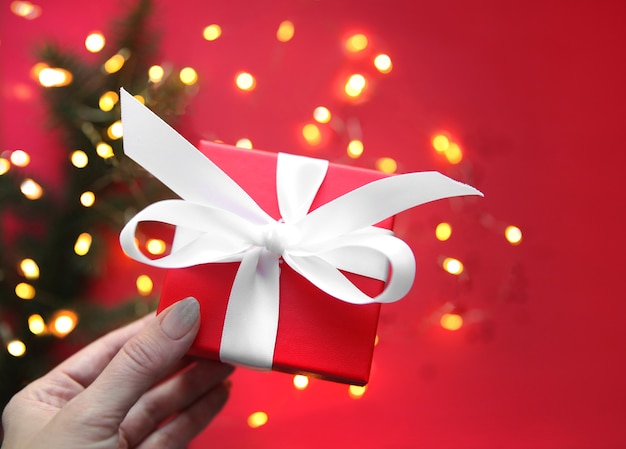 Red gift box in female hands, decorated with a white ribbon on a red background